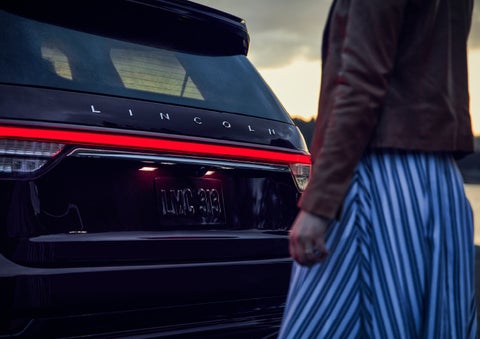 A person is shown near the rear of a 2024 Lincoln Aviator® SUV as the Lincoln Embrace illuminates the rear lights | Dave Sinclair Lincoln St. Peters in Saint Peters MO